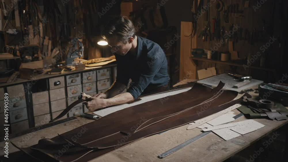 Young professional tailor making leather belts at his workshop, creating handmade original accessories, cutting material