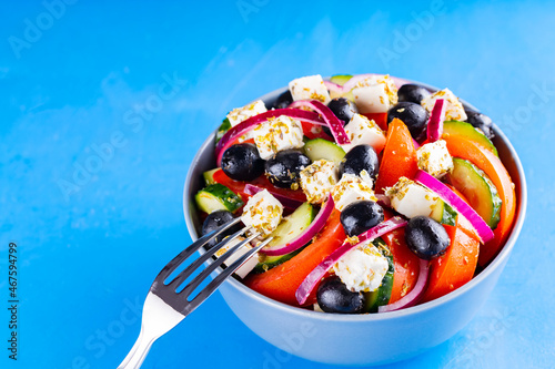 Greek salad and fork on a blue background. Fresh vegetables, feta cheese and black olives. Trendy food. Copy space. Top view