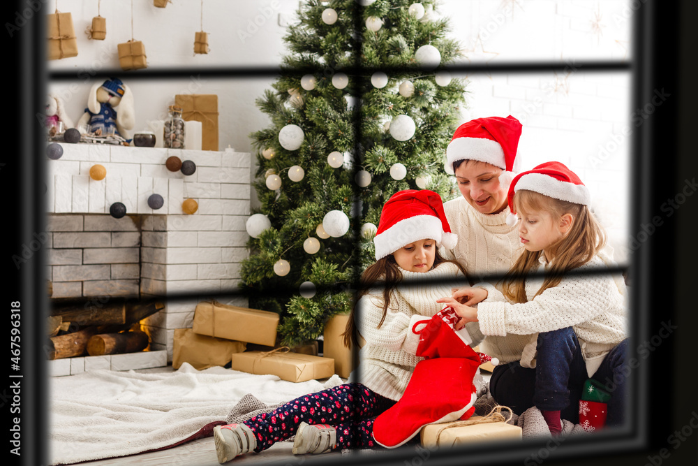 Fototapeta premium Young big family celebrating Christmas enjoying dinner, view from outside through a window into a decorated living room with tree and candle lights, happy parents eating with three kids.