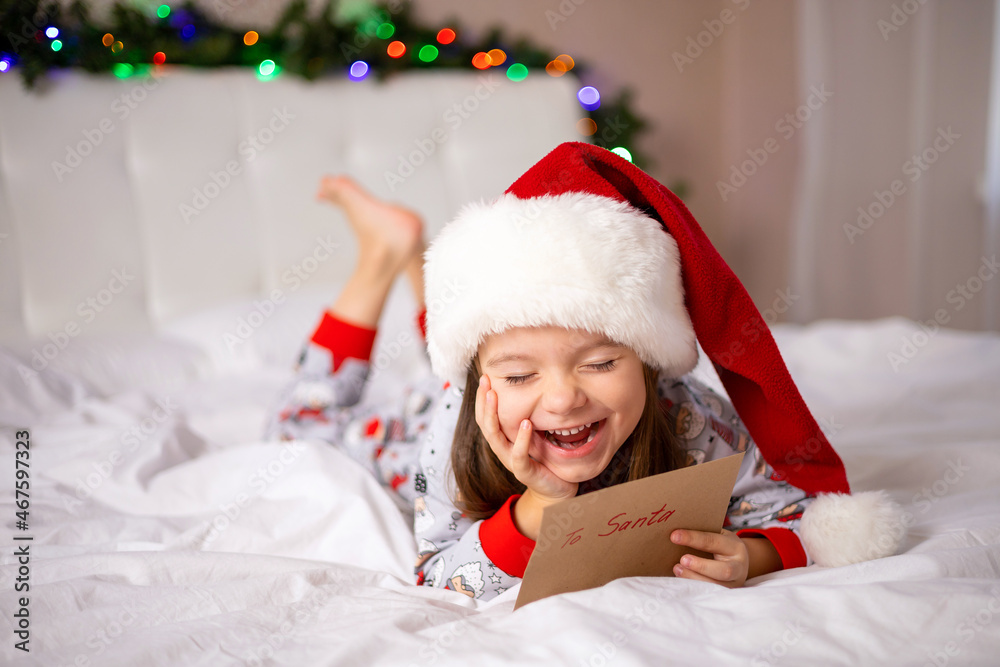 Fototapeta premium Merry Christmas and Happy Holidays. The little happy girl on the bed wrote a letter to Santa Claus and holds the envelope in her hand.