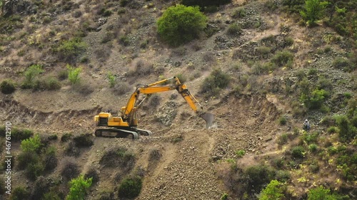 Bright yellow excavator digs rock on mountain ridge. Road works in hills, yellow sandy land. Construction work in highlands. Heavy machinery at industrial construction site. Aerial drone shot zoom in