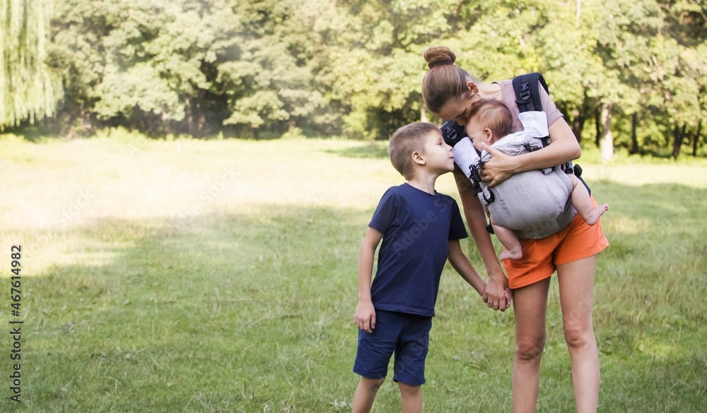 Fototapeta premium Mother and two children are walking in the park. Family walk. Mom carries a newborn in a sling and holds the hand of an older child