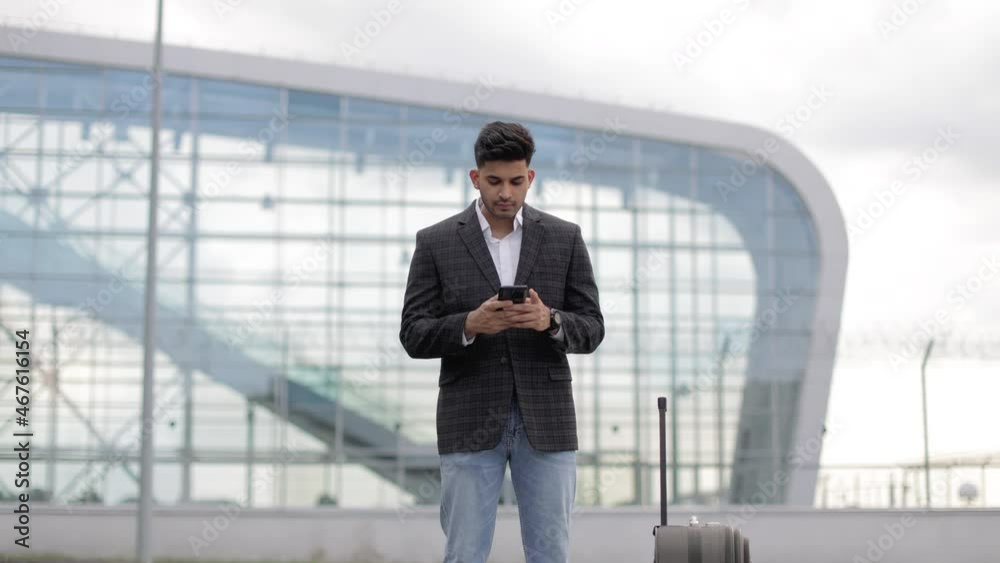 Young Arabian man standing outside in front of airport gate and checking his flight schedule on phone. Young man browsing on cellphone, typing message. Urban, travel and communication concept Young Arabian man standing outside in front of airport gate and checking his flight schedule on phone. Young man browsing on cellphone, typing message. Urban, travel and communication concept