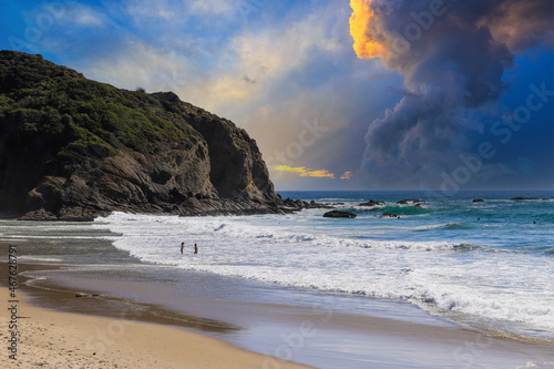 a stunning shot of the vast blue ocean water and waves rolling to the beach and crashing into the rocks with people walking along the beach with powerful clouds in a blue sky at Dana Strands Beach