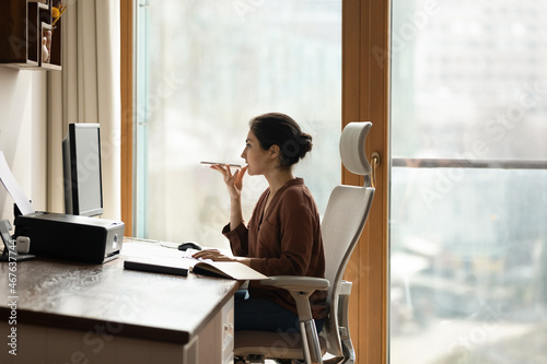 Serious young Indian businesswoman sit on chair at workplace holds smartphone talk to client on speaker phone, leaves voicemail, voice message communicates remotely use modern tech, connection concept photo