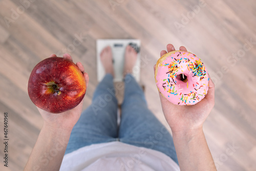 A woman stands on the scales, holds a donut and an apple in her hands, top view.