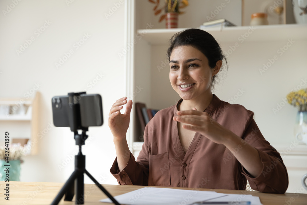 Young Indian woman put cellphone on tripod share knowledge record video vlog seated at table. Influencing, job interview remotely, modern technologies usage, educational webinar and e-coaching concept Young Indian woman put cellphone on tripod share knowledge record video vlog seated at table. Influencing, job interview remotely, modern technologies usage, educational webinar and e-coaching concept