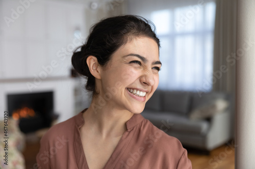 Laughing face of young Indian woman close up head shot portrait. Cheerful female housewife standing alone in modern coy living room looks aside feels overjoyed giggling over joke, humour, fun concept