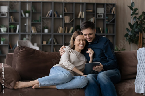 Happy couple hugging, using tablet together, relaxing on couch, smiling wife and husband looking at electronic device screen, watching funny video or chatting online, spending leisure time at home