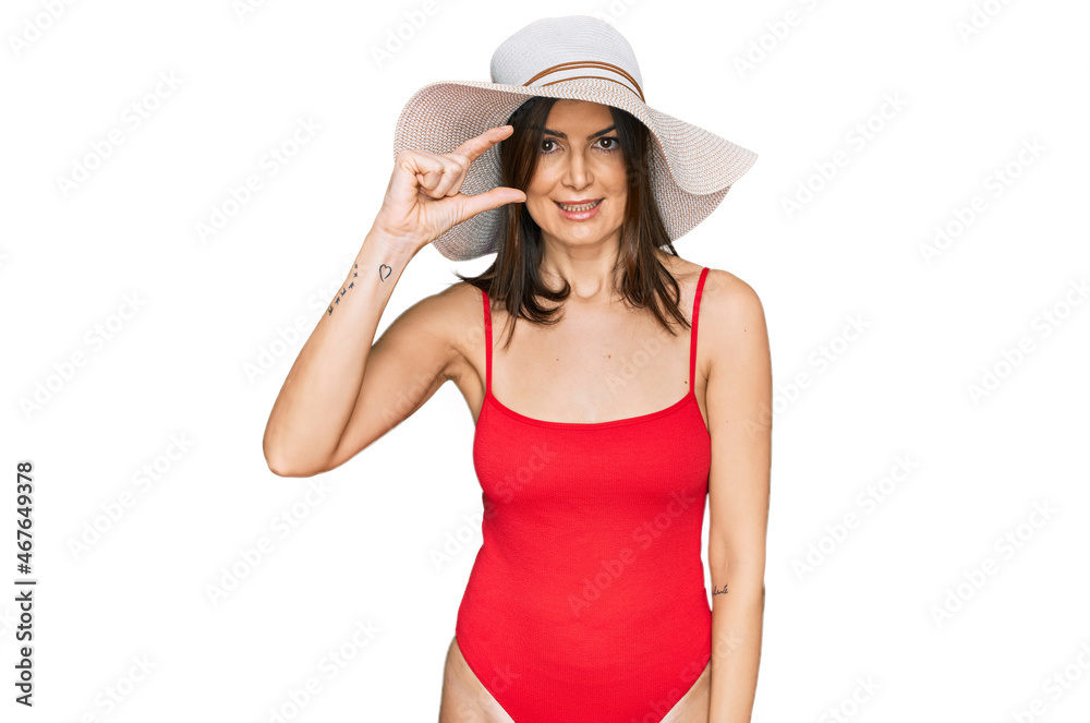 Beautiful brunette woman wearing swimsuit and summer hat smiling and confident gesturing with hand doing small size sign with fingers looking and the camera. measure concept. Beautiful brunette woman wearing swimsuit and summer hat smiling and confident gesturing with hand doing small size sign with fingers looking and the camera. measure concept.