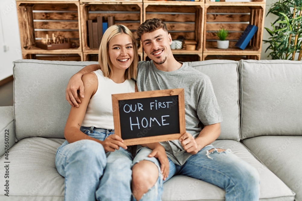 Young caucasian couple smiling happy holding blackboard with our first home message sitting on the sofa. - obrazy, fototapety, plakaty 