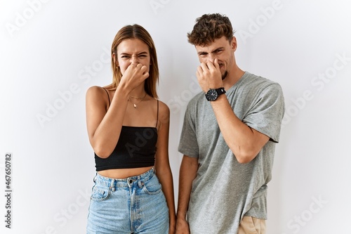 Young beautiful couple standing together over isolated background smelling something stinky and disgusting, intolerable smell, holding breath with fingers on nose. bad smell