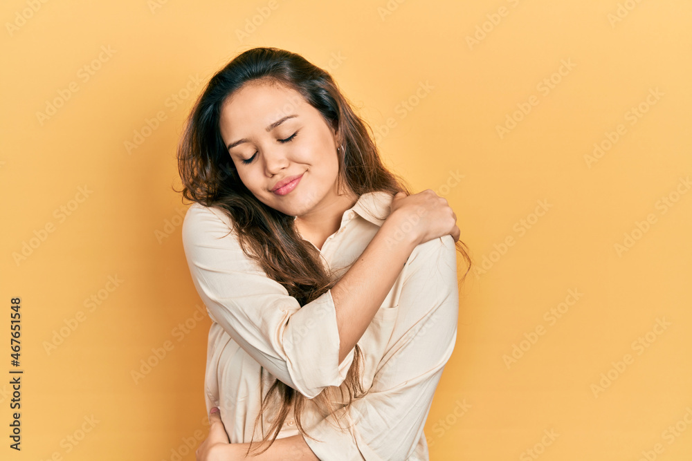 Fototapeta premium Young hispanic girl wearing casual clothes hugging oneself happy and positive, smiling confident. self love and self care