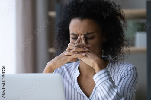 Close up unhappy exhausted African American businesswoman feeling pain and dizziness, overworked young female touching nose bridge, suffering from strong headache or migraine after long laptop using