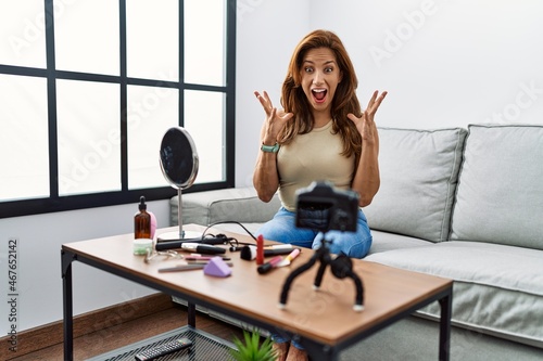 Middle age hispanic woman recording make up tutorial with smartphone at home celebrating victory with happy smile and winner expression with raised hands