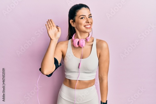 Young hispanic woman wearing gym clothes and using headphones waiving saying hello happy and smiling, friendly welcome gesture