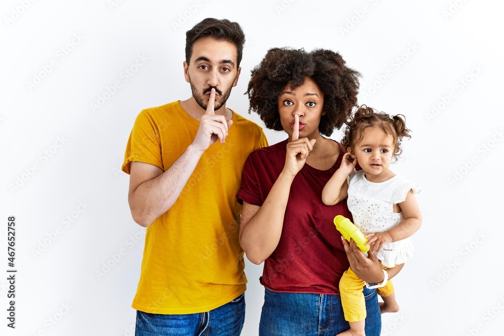 Interracial young family of black mother and hispanic father with daughter asking to be quiet with finger on lips. silence and secret concept.