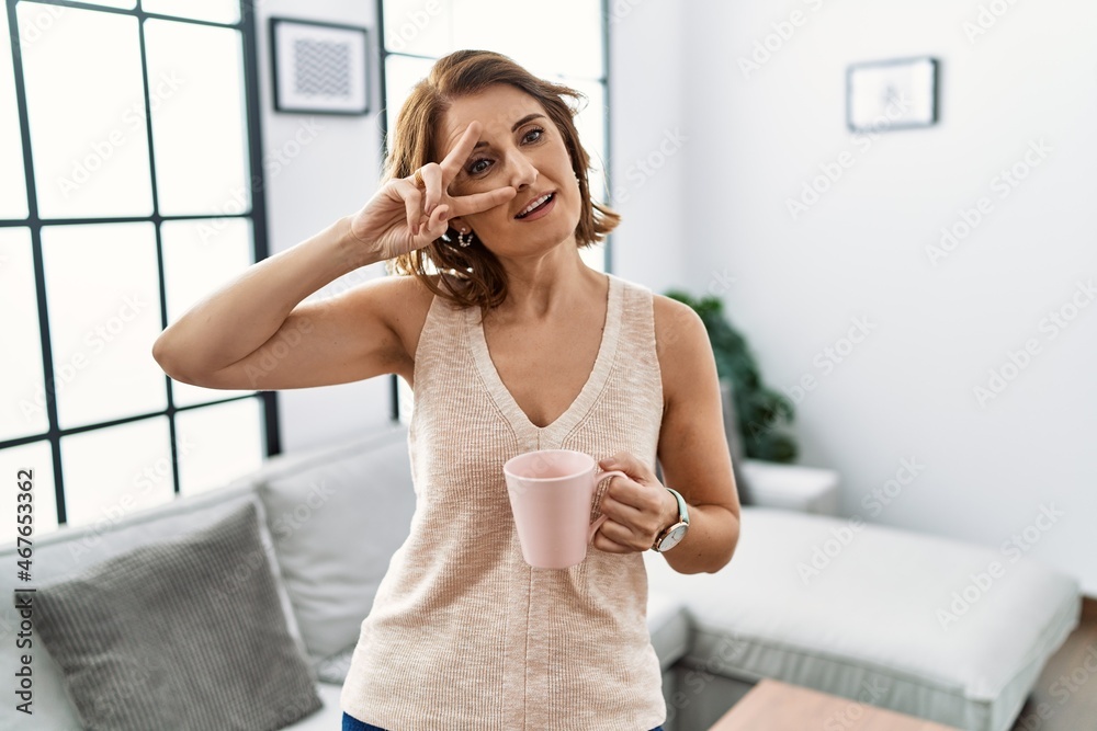 Middle age woman drinking a cup coffee at home doing peace symbol with fingers over face, smiling cheerful showing victory Middle age woman drinking a cup coffee at home doing peace symbol with fingers over face, smiling cheerful showing victory