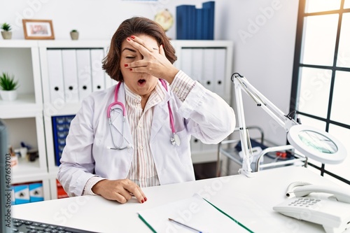 Middle age hispanic woman wearing doctor uniform and stethoscope at the clinic peeking in shock covering face and eyes with hand  looking through fingers with embarrassed expression.