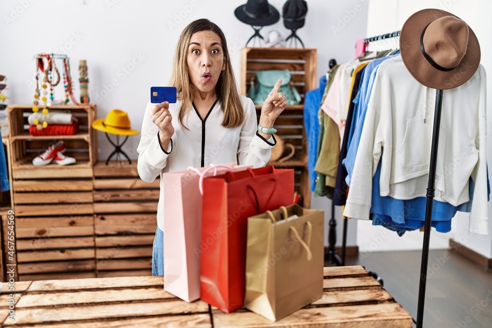 Young hispanic woman holding shopping bags and credit card at clothing store surprised pointing with finger to the side, open mouth amazed expression. Young hispanic woman holding shopping bags and credit card at clothing store surprised pointing with finger to the side, open mouth amazed expression.