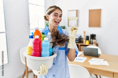 Young blonde woman wearing cleaner uniform holding cleaning products with a big smile on face, pointing with hand finger to the side looking at the camera.