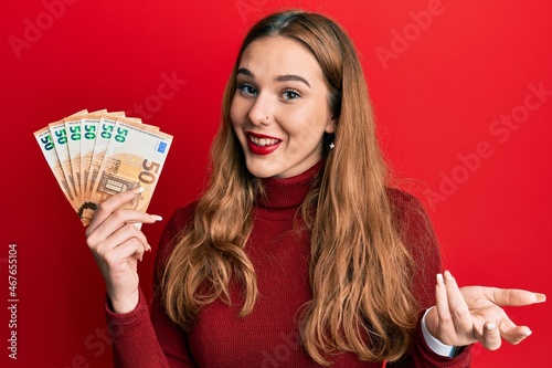 Young blonde woman holding euro banknotes celebrating achievement with happy smile and winner expression with raised hand