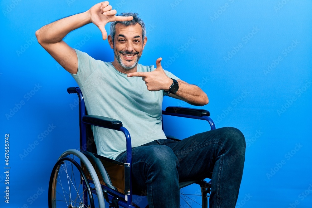 Obraz premium Handsome middle age man with grey hair sitting on wheelchair smiling making frame with hands and fingers with happy face. creativity and photography concept.