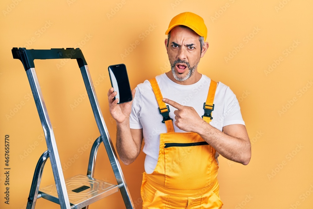 Handsome middle age man with grey hair standing by ladder showing smartphone in shock face, looking skeptical and sarcastic, surprised with open mouth Handsome middle age man with grey hair standing by ladder showing smartphone in shock face, looking skeptical and sarcastic, surprised with open mouth