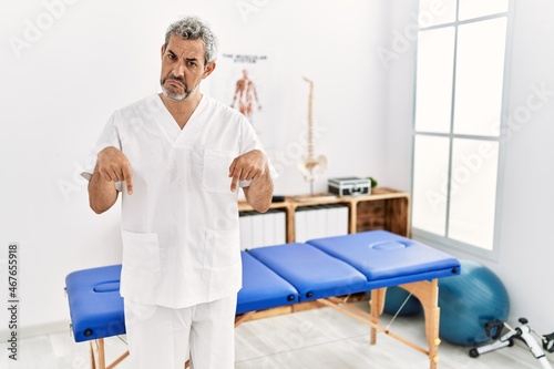 Middle age hispanic therapist man working at pain recovery clinic pointing down looking sad and upset  indicating direction with fingers  unhappy and depressed.