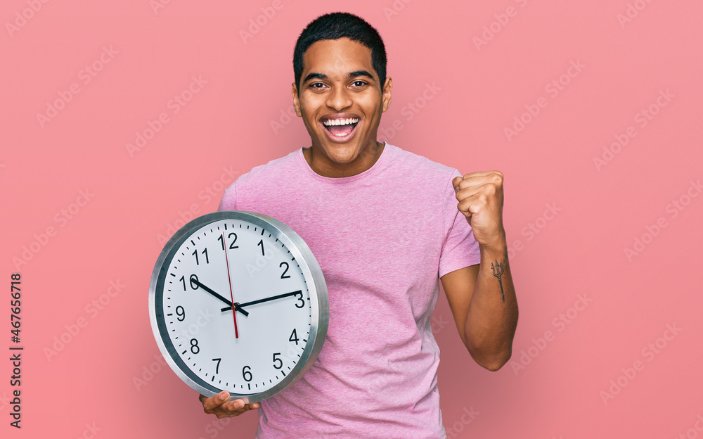 Young handsome hispanic man holding big clock screaming proud, celebrating victory and success very excited with raised arms Young handsome hispanic man holding big clock screaming proud, celebrating victory and success very excited with raised arms