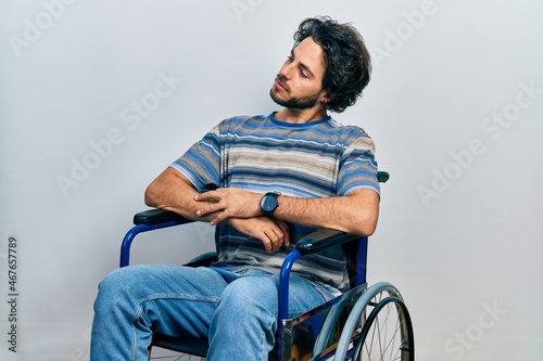 Handsome hispanic man sitting on wheelchair looking to the side with arms crossed convinced and confident