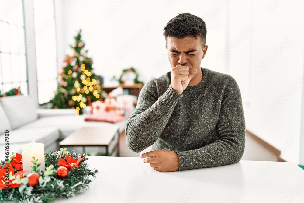 Young hispanic man sitting on the table by christmas tree feeling unwell and coughing as symptom for cold or bronchitis. health care concept.