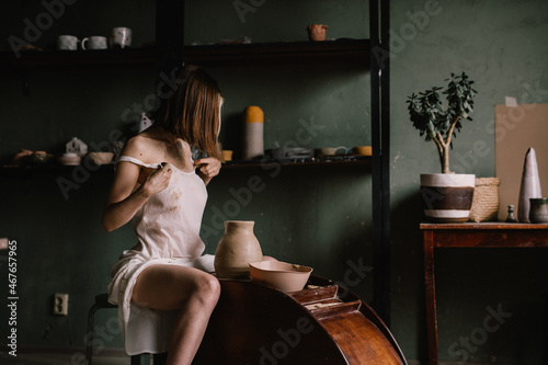 talented sculptor girl in thin light negligee sculpts a jug of clay in pottery workshop near large window. Concept art of sculpture