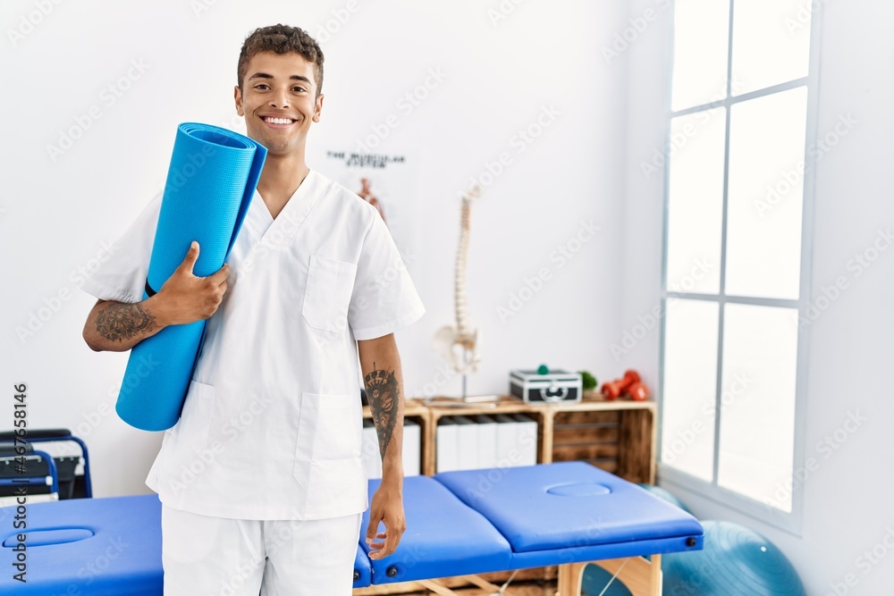 Young hispanic man working as physiotherapist holding yoga mat at physiotherapy room