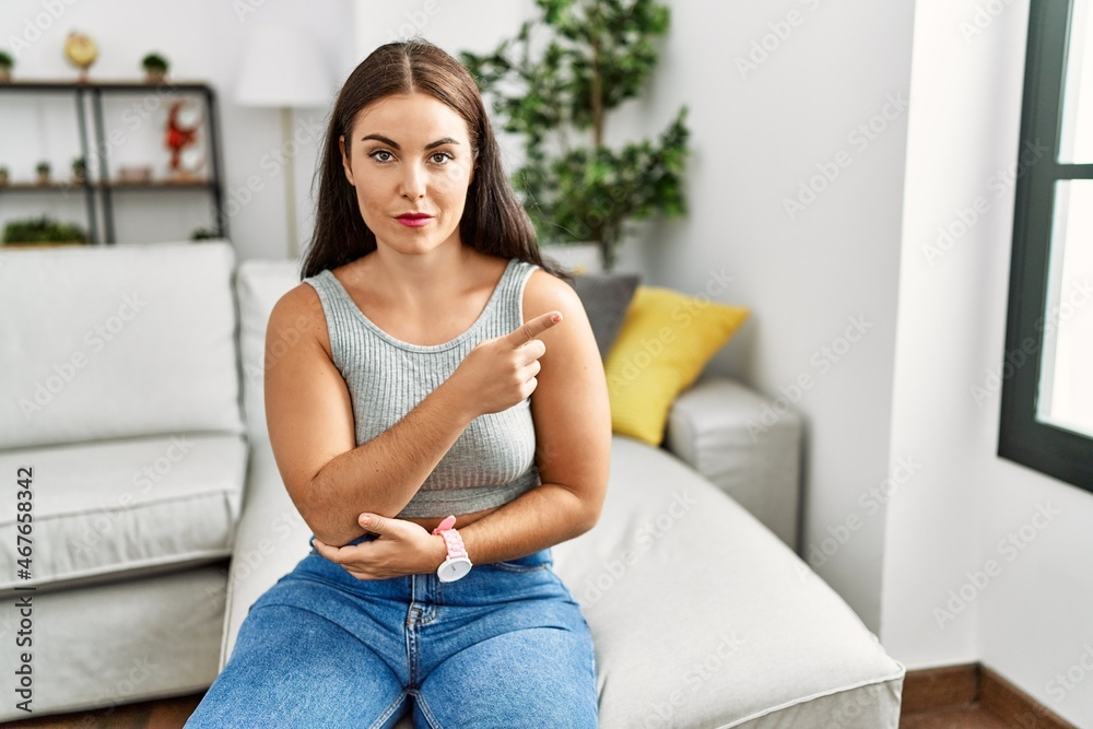 Young brunette woman sitting on the sofa at home pointing with hand finger to the side showing advertisement, serious and calm face Young brunette woman sitting on the sofa at home pointing with hand finger to the side showing advertisement, serious and calm face