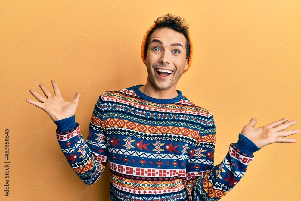 Young handsome man wearing wool hat and colorful sweater celebrating victory with happy smile and winner expression with raised hands Young handsome man wearing wool hat and colorful sweater celebrating victory with happy smile and winner expression with raised hands