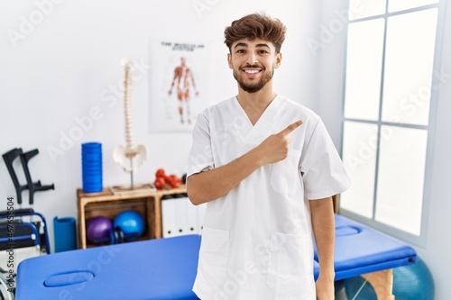 Young arab man working at pain recovery clinic cheerful with a smile of face pointing with hand and finger up to the side with happy and natural expression on face