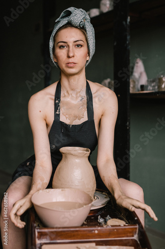 young sculptor, a girl with bandage on her head and in black apron, sculpts a clay product in pottery workshop near large window. Concept hobby sculpture