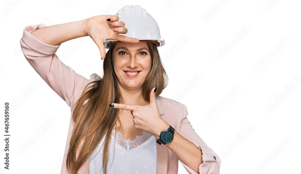 Young blonde woman wearing architect hardhat smiling making frame with hands and fingers with happy face. creativity and photography concept. Young blonde woman wearing architect hardhat smiling making frame with hands and fingers with happy face. creativity and photography concept.