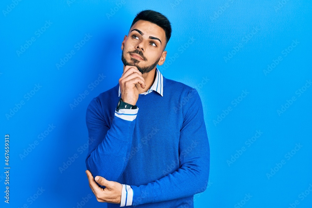 Young hispanic man with beard wearing casual blue sweater with hand on chin thinking about question, pensive expression. smiling and thoughtful face. doubt concept. Young hispanic man with beard wearing casual blue sweater with hand on chin thinking about question, pensive expression. smiling and thoughtful face. doubt concept.