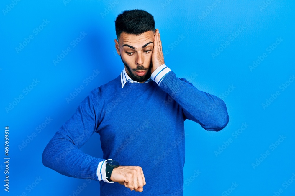 Young hispanic man with beard wearing casual blue sweater looking at the watch time worried, afraid of getting late Young hispanic man with beard wearing casual blue sweater looking at the watch time worried, afraid of getting late