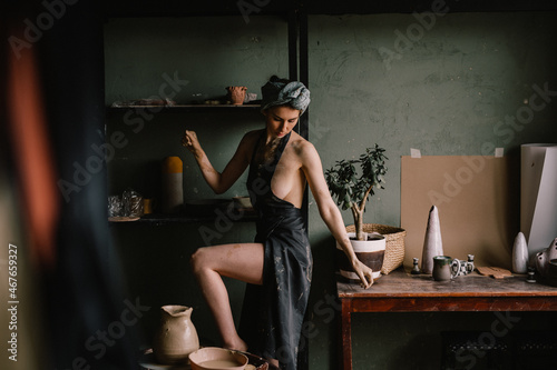 beautiful sculptor girl with bandage on her head and a black apron on her naked body is standing in pottery workshop near large window. concept is beauty and art.