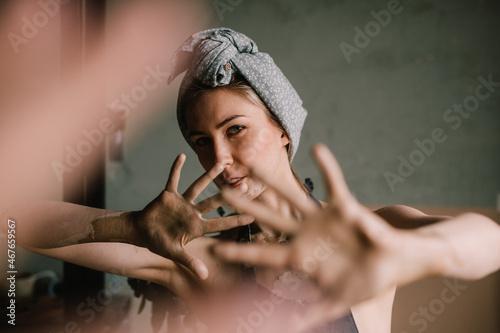 beautiful sculptor girl with a headband and a black apron shows her hands stained with clay. camera focus is on girl's face, blur is on her hands.
