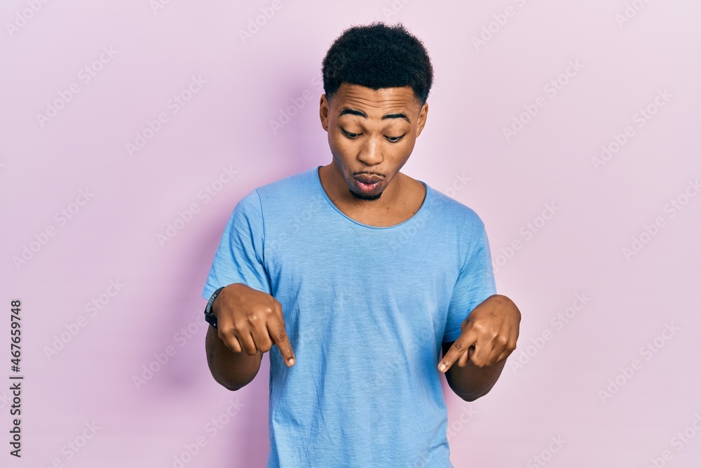 Young african american man wearing casual blue t shirt pointing down with fingers showing advertisement, surprised face and open mouth Young african american man wearing casual blue t shirt pointing down with fingers showing advertisement, surprised face and open mouth
