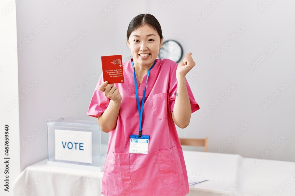 Young asian nurse woman at political campaign election holding swiss passport screaming proud, celebrating victory and success very excited with raised arm Young asian nurse woman at political campaign election holding swiss passport screaming proud, celebrating victory and success very excited with raised arm