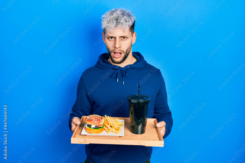 Young hispanic man with modern dyed hair eating a tasty classic burger with fries and soda in shock face, looking skeptical and sarcastic, surprised with open mouth Young hispanic man with modern dyed hair eating a tasty classic burger with fries and soda in shock face, looking skeptical and sarcastic, surprised with open mouth