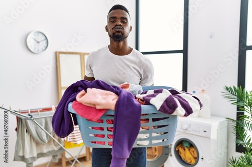 Young african man holding laundry basket puffing cheeks with funny face. mouth inflated with air, catching air.