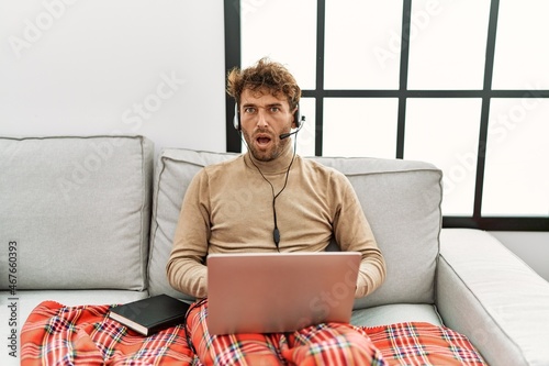 Young handsome man with beard wearing operator headset working from home in shock face, looking skeptical and sarcastic, surprised with open mouth