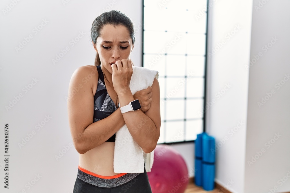 Young brunette woman wearing sportswear and towel at the gym looking stressed and nervous with hands on mouth biting nails. anxiety problem.