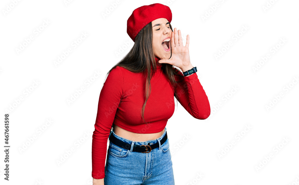 Young brunette teenager wearing french look with beret shouting and screaming loud to side with hand on mouth. communication concept. Young brunette teenager wearing french look with beret shouting and screaming loud to side with hand on mouth. communication concept.